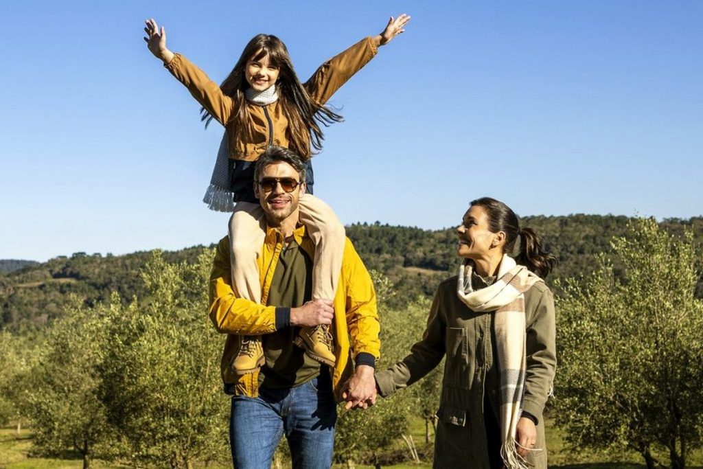 Família ao ar livre com criança nos ombros do pai entre oliveiras em dia ensolarado
