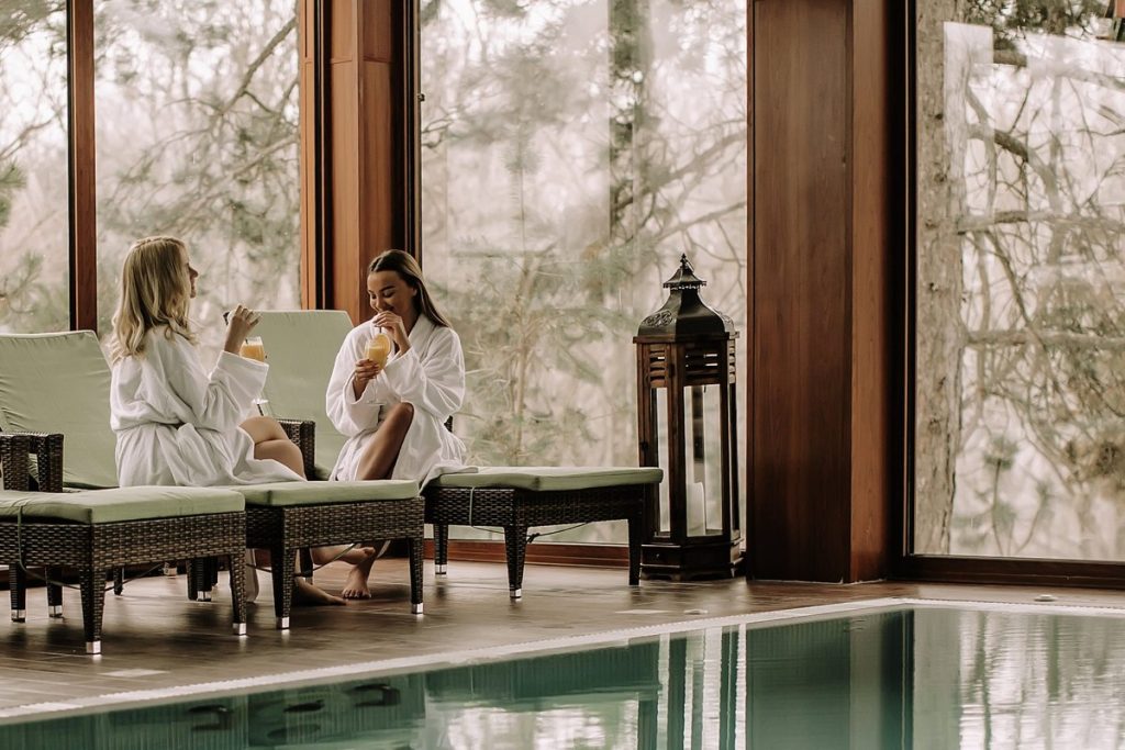 Duas mulheres em roupões brancos relaxando em spa com piscina coberta e vista para a floresta